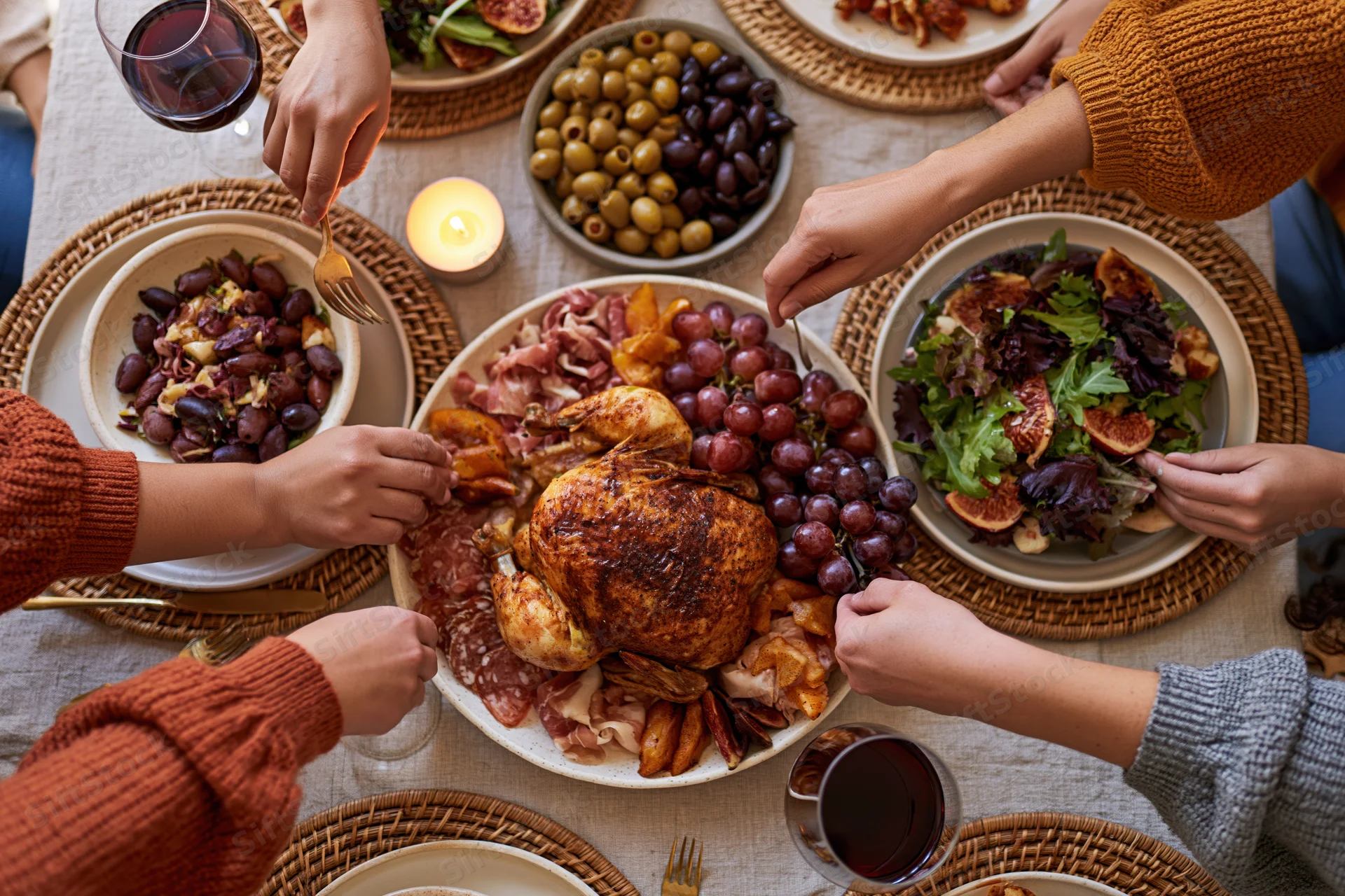 Overhead view of hands reaching for food at a holiday dinner table with roasted chicken, grapes, salad, and wine