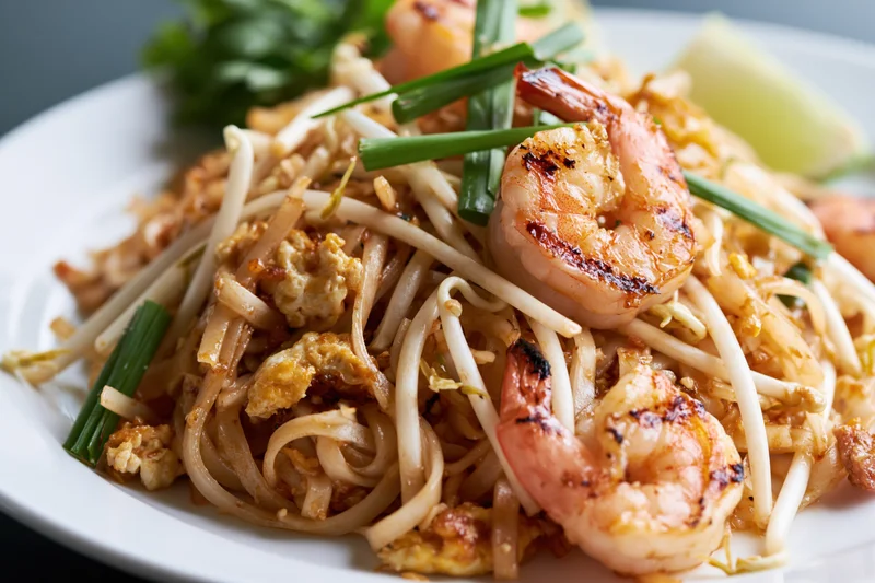 Pad thai noodles with grilled shrimp and scrambled egg on a white plate garnished with bean sprouts and chives