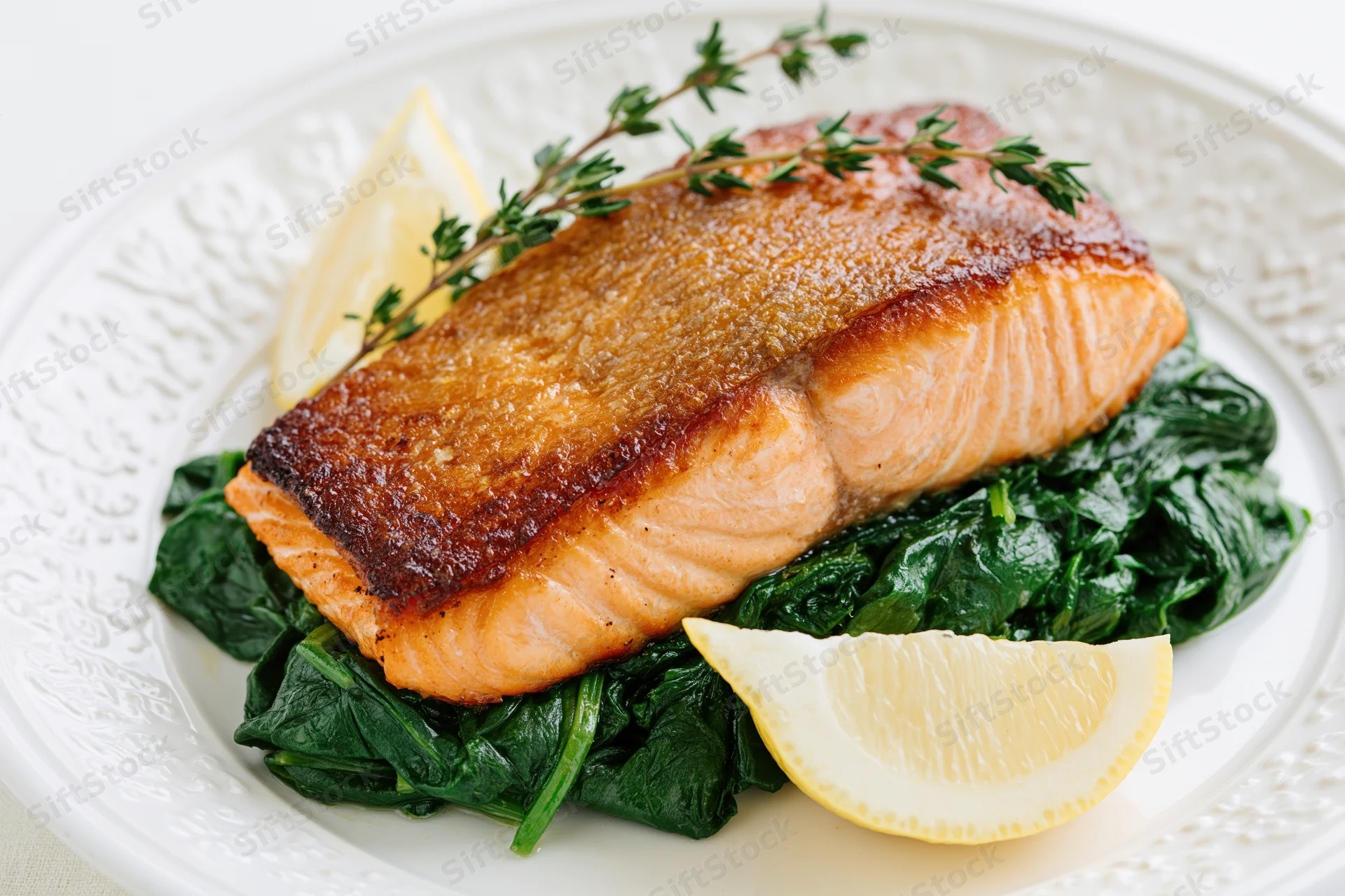 Pan-seared salmon fillet with crispy skin on a bed of spinach and lemon wedges on a white decorative plate