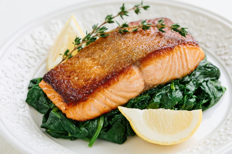 Pan-seared salmon fillet with crispy skin on a bed of spinach and lemon wedges on a white decorative plate
