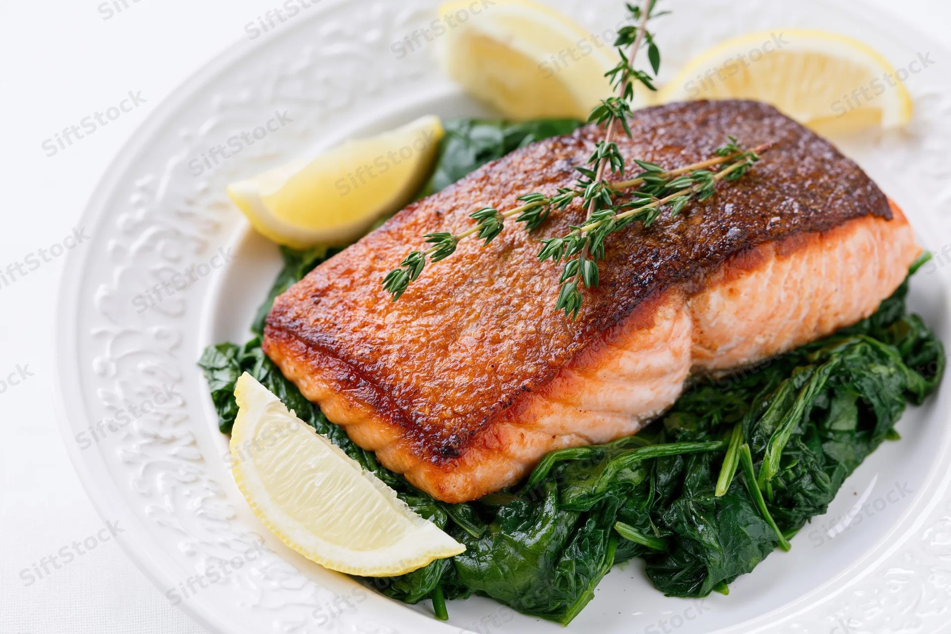 Pan-seared salmon fillet with crispy skin on a bed of spinach garnished with lemon wedges and thyme on a white decorative plate