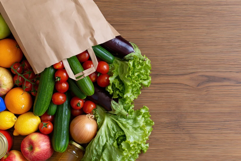 Paper grocery bag tipped on its side spilling fresh vegetables and fruits onto wooden table