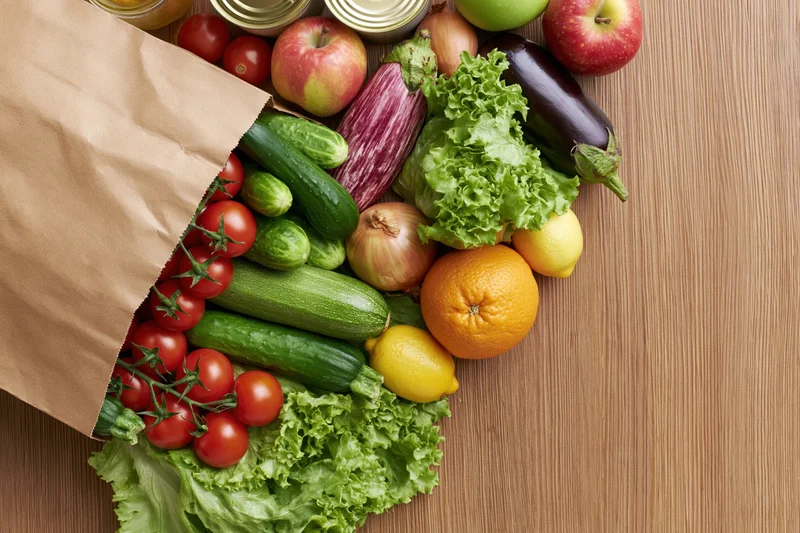 Paper grocery bag tipped on its side spilling fresh vegetables and fruits onto wooden table