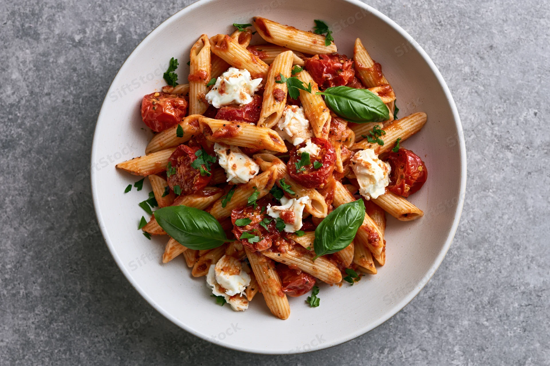 Penne pasta with roasted cherry tomatoes and torn mozzarella in a white bowl on gray surface