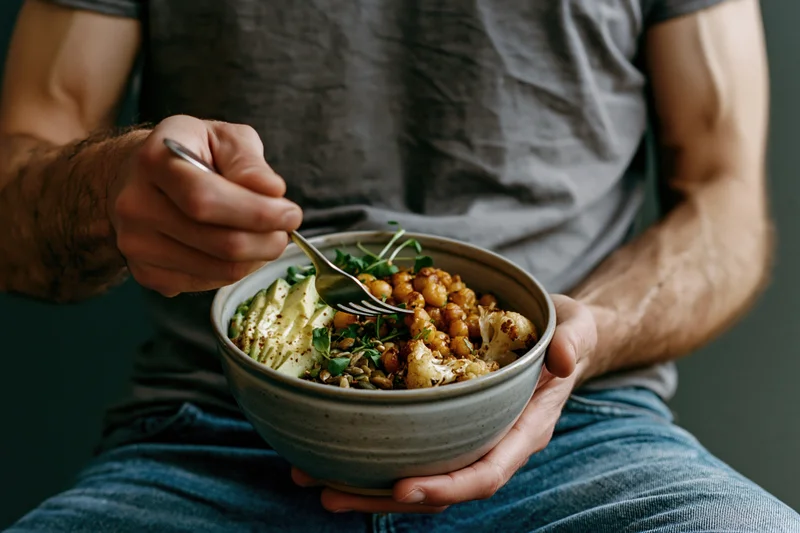 Man holding ceramic bowl with vegan grain bowl, eating with fork, seated on lap