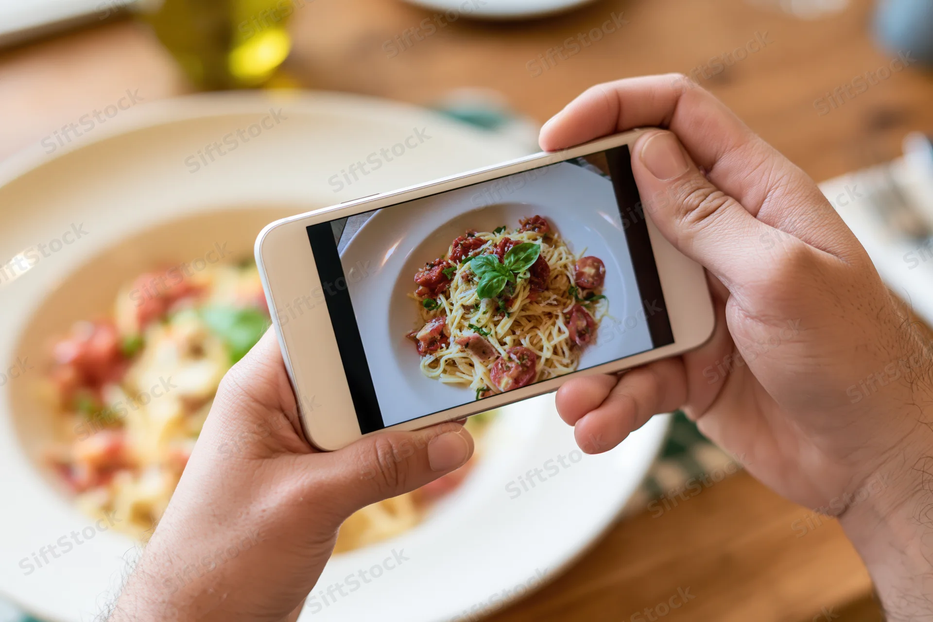 Hands holding smartphone photographing plate of spaghetti with tomato sauce and basil on wooden table