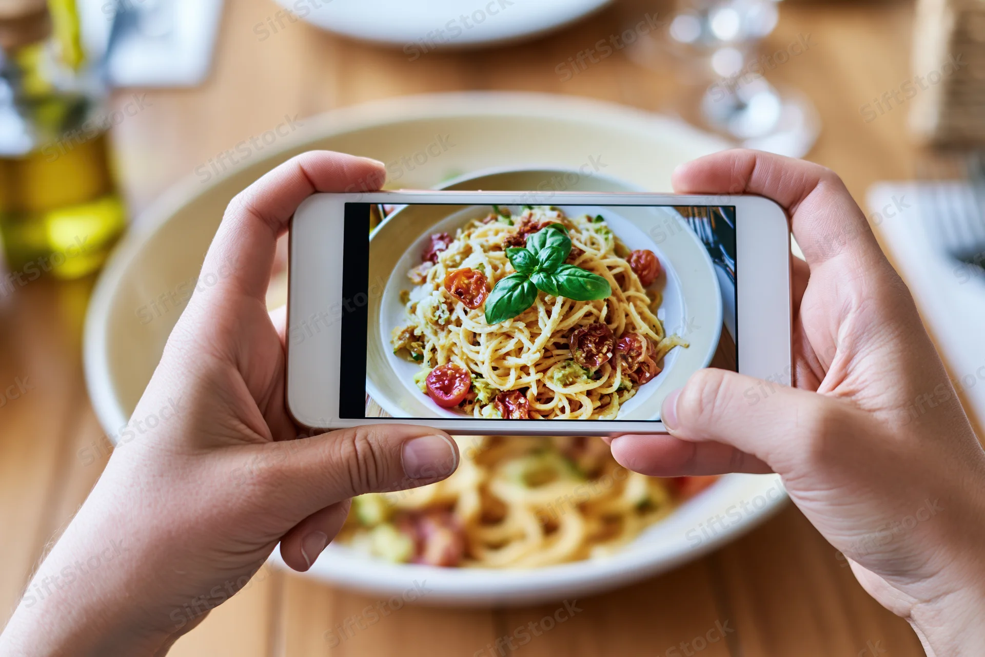 Hands holding smartphone photographing bowl of spaghetti with basil and tomatoes on wooden table