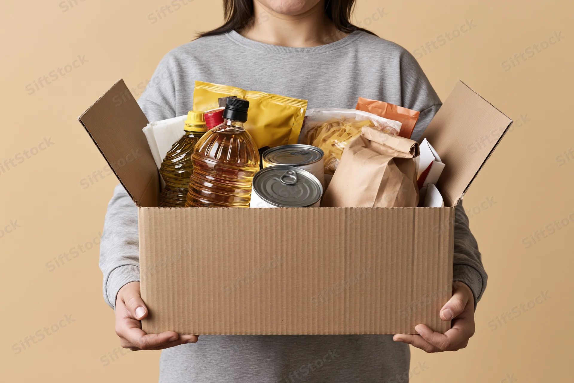 Woman holding open cardboard box filled with pantry staples against beige background