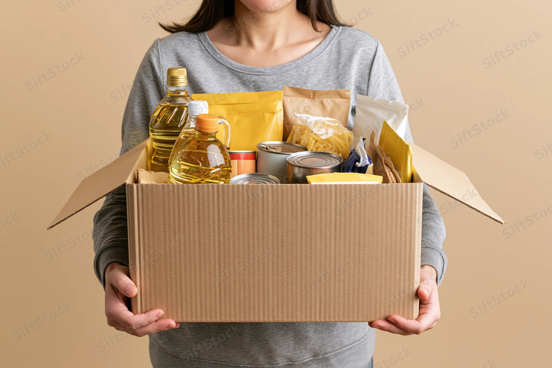 Woman holding open cardboard box filled with pantry staples against neutral background
