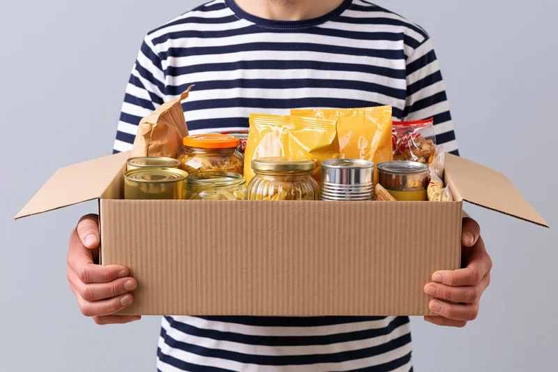 Man in striped shirt holding open cardboard box filled with canned and packaged food items