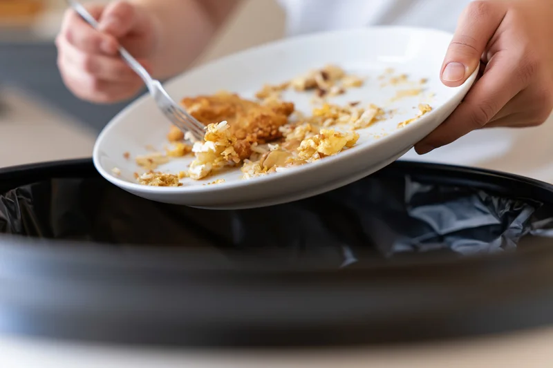 Person scraping leftover food scraps off a white plate into a trash bin