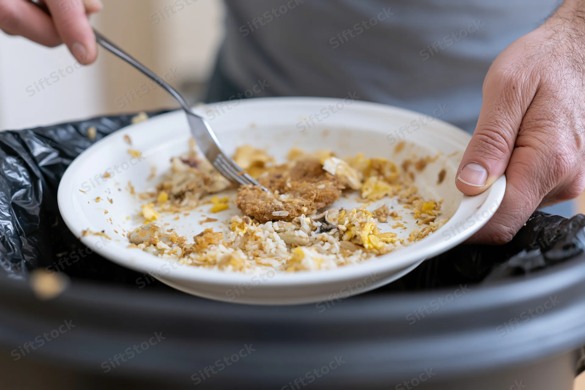 Man scraping leftover food scraps from a white plate into a trash bin