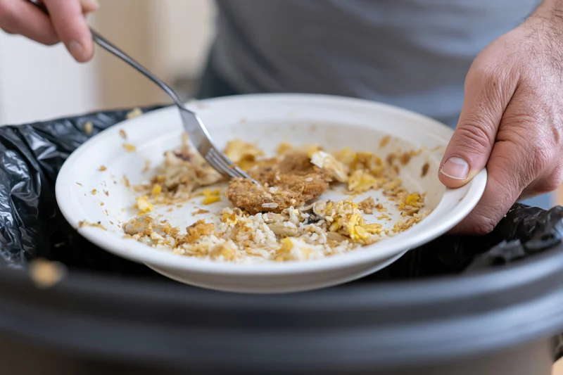 Man scraping leftover food scraps from a white plate into a trash bin