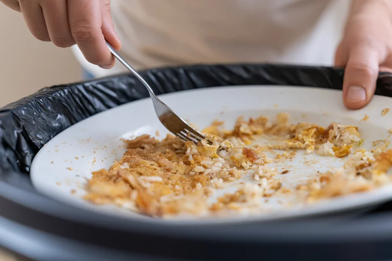 Person scraping food scraps from a white plate into a trash bin