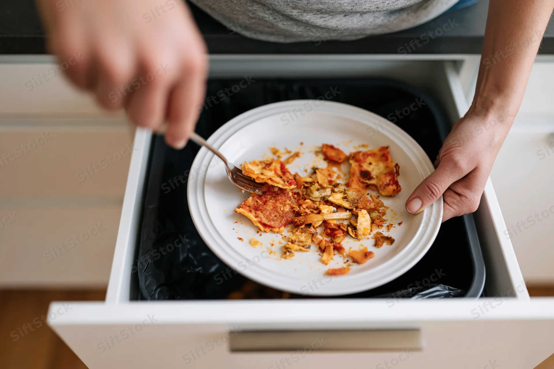 Person scraping pizza scraps from plate into kitchen drawer trash bin