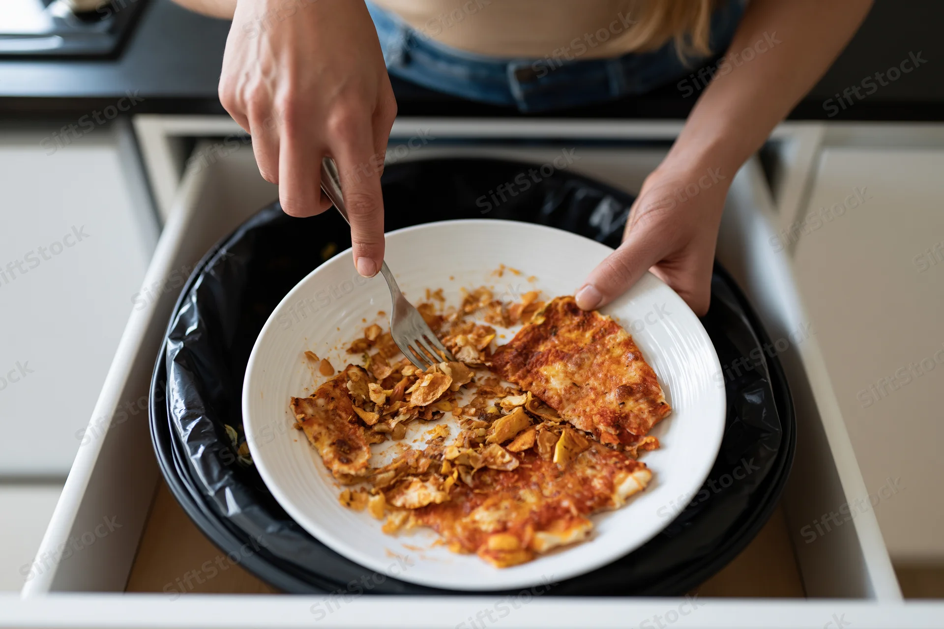 Woman scraping leftover pizza and food scraps from a plate into a kitchen trash bin drawer