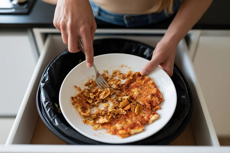 Woman scraping leftover pizza and food scraps from a plate into a kitchen trash bin drawer