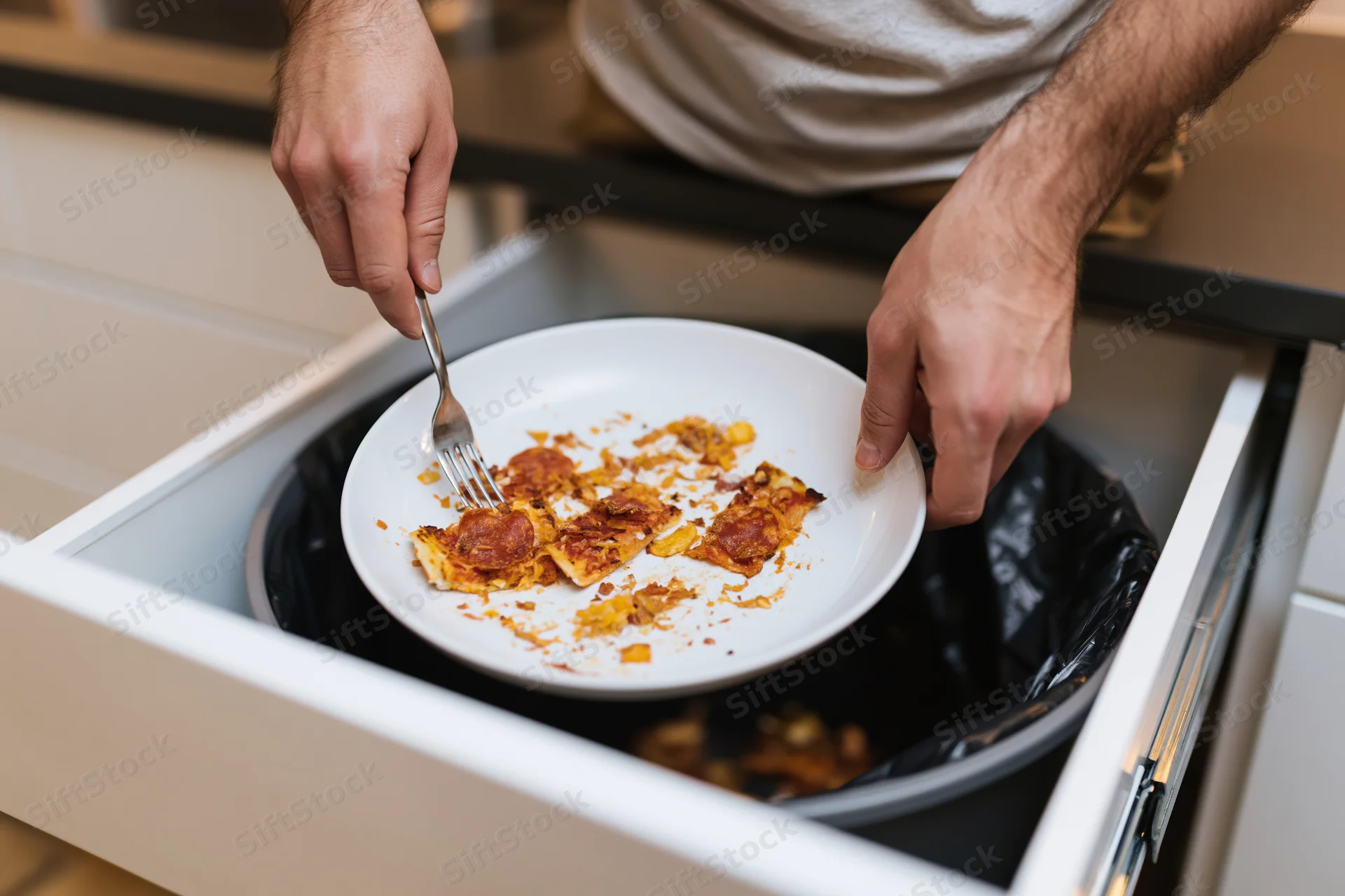 Man scraping leftover pizza scraps from a plate into a kitchen drawer trash bin