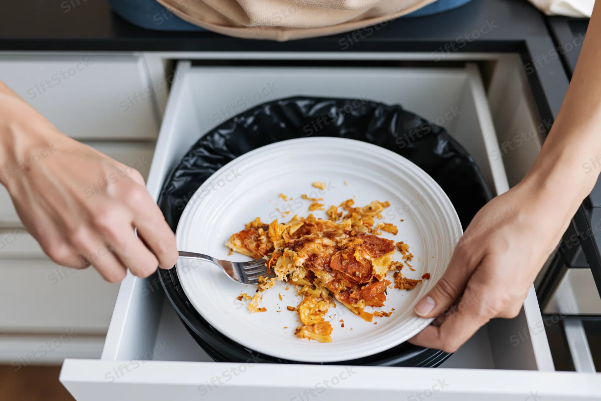 Hands scraping pizza scraps from a plate into a kitchen drawer trash bin