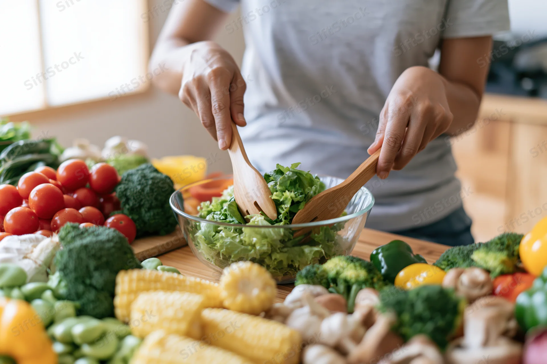 Woman tossing a green salad in a glass bowl surrounded by fresh vegetables on a kitchen countertop