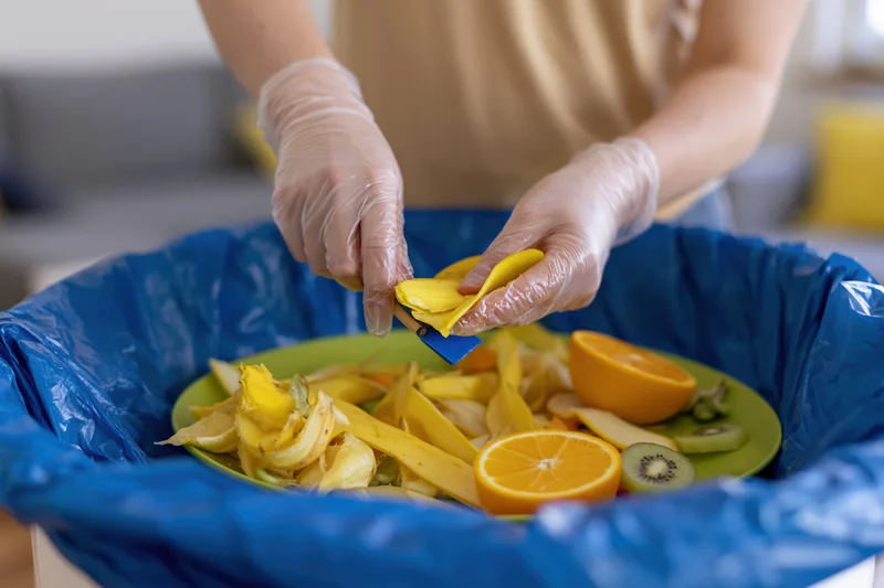 Woman in plastic gloves sorting fruit peels into a compost bin in a kitchen