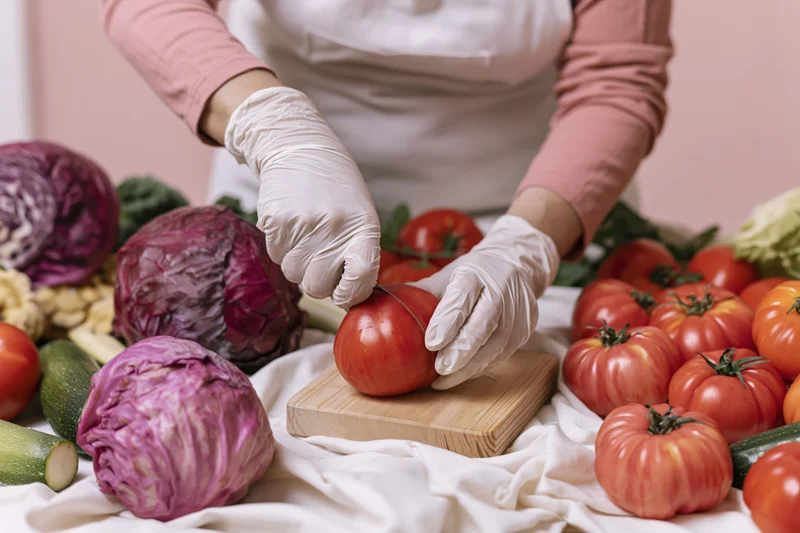 Woman slicing a ripe tomato on a wooden cutting board surrounded by fresh vegetables in a kitchen setting