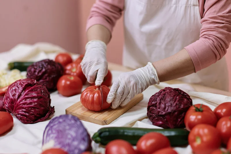 Woman slicing a tomato on a wooden cutting board surrounded by fresh vegetables on a kitchen table
