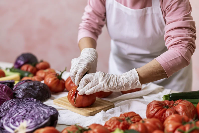 Woman slicing a ripe tomato on a wooden cutting board surrounded by fresh vegetables in a kitchen setting