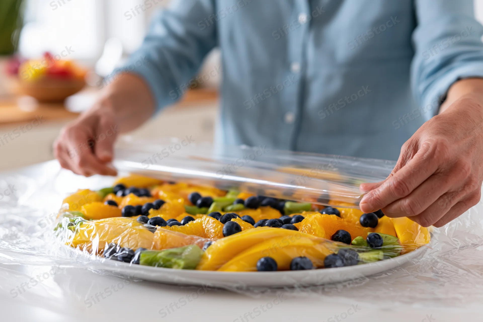 Man wrapping a round fruit platter with plastic cling wrap on a kitchen countertop