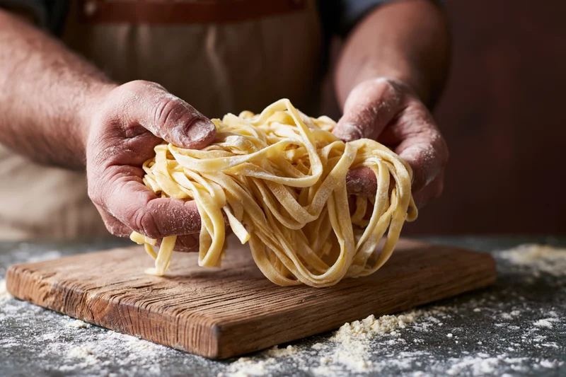 Flour-dusted hands lifting a nest of fresh tagliatelle over a wooden cutting board