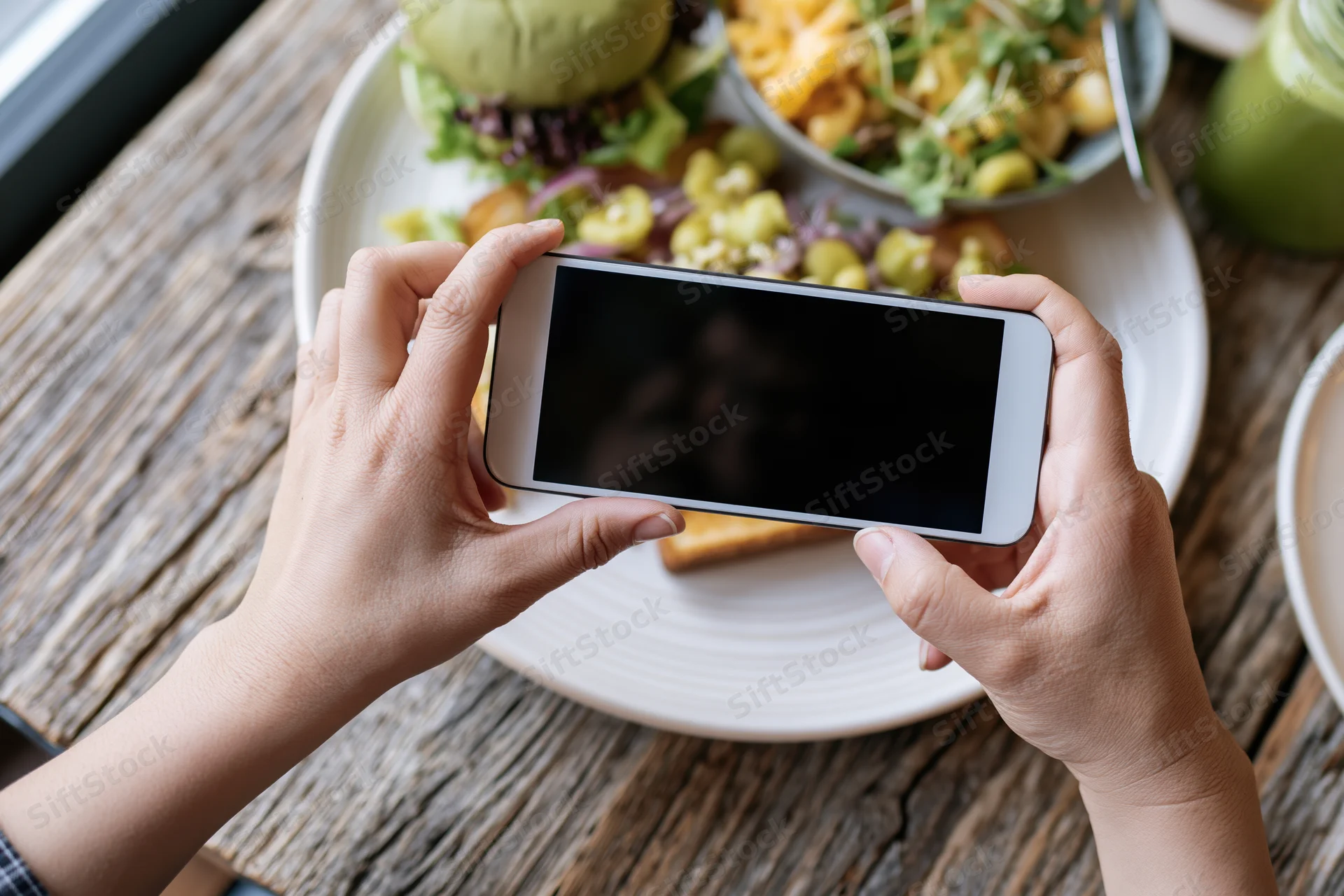 Hands holding smartphone horizontally photographing a plated meal on a wooden table