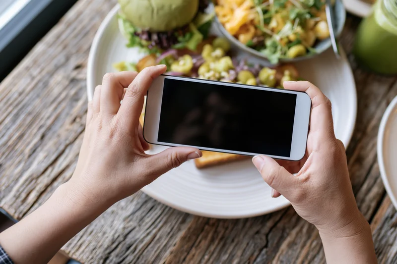 Hands holding smartphone horizontally photographing a plated meal on a wooden table