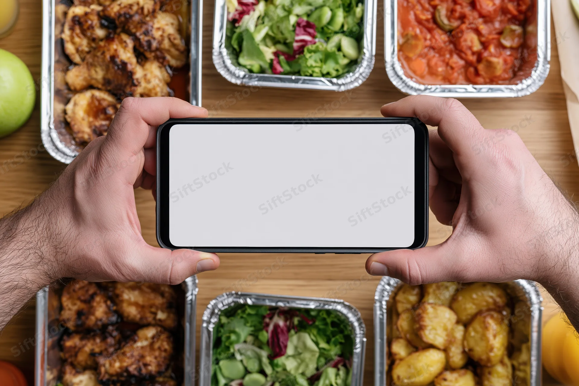 Hands holding smartphone with blank white screen over meal containers on wooden table