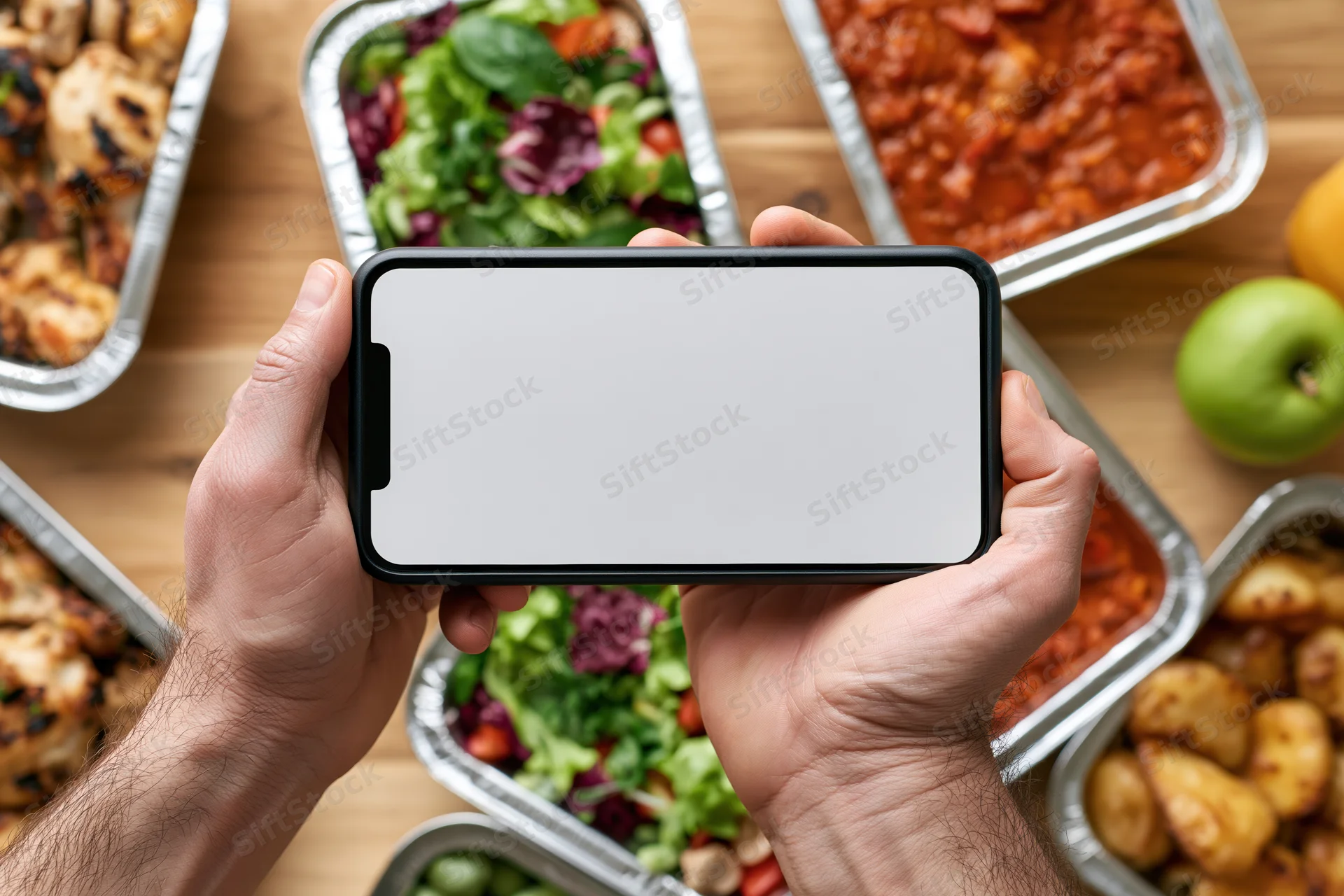 Hands holding smartphone with blank white screen over meal containers on wooden table