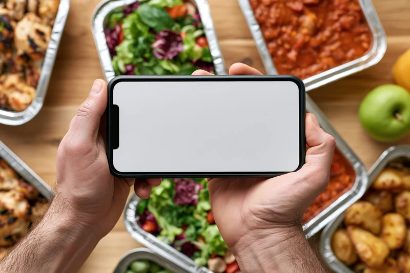 Hands holding smartphone with blank white screen over meal containers on wooden table