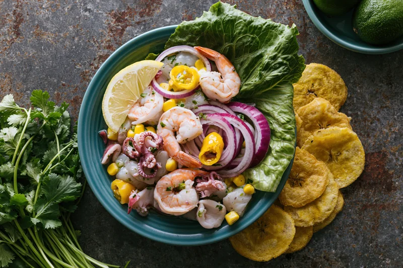 Peruvian seafood ceviche mixto in a teal ceramic bowl with lemon, onion, pepper, lettuce, cilantro, and fried plantain on a dark textured surface