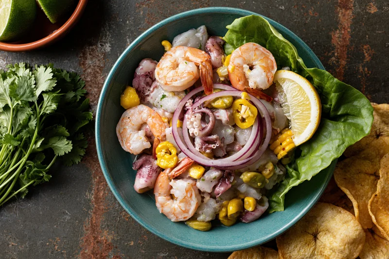 Peruvian seafood ceviche mixto in a teal ceramic bowl with shrimp, squid, red onion, lemon, corn, chili, and cilantro, served with plantain chips on a rustic surface