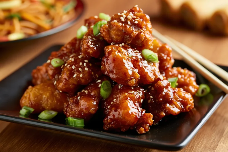 Crispy glazed orange chicken pieces piled high on a black square plate with sesame seeds and scallions, chopsticks beside, on wooden table