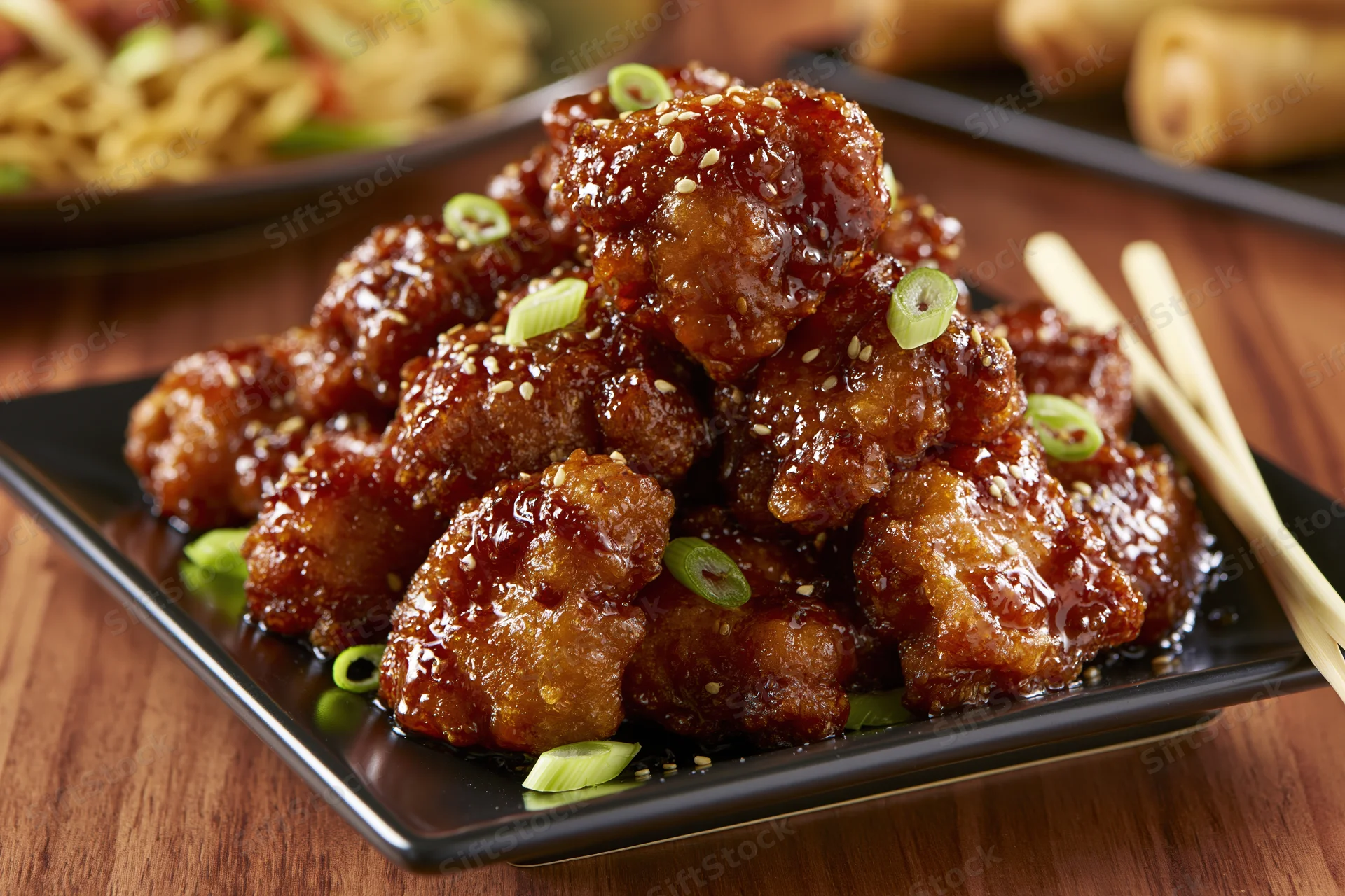 Crispy glazed orange chicken pieces piled high on a black plate with sesame seeds and scallions, served with chopsticks on a wooden table