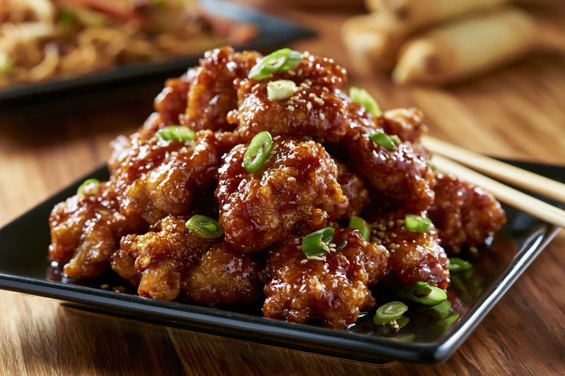 Crispy glazed orange chicken pieces piled high on a black square plate with sesame seeds and scallions, chopsticks resting beside, on a wooden table