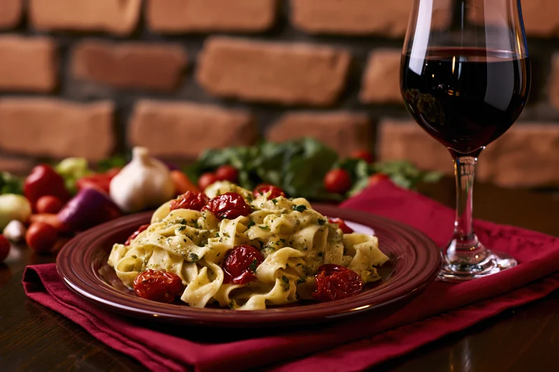 Plate of tagliatelle pasta with cherry tomatoes and herbs on a red napkin beside a glass of red wine on a wooden table with brick wall background