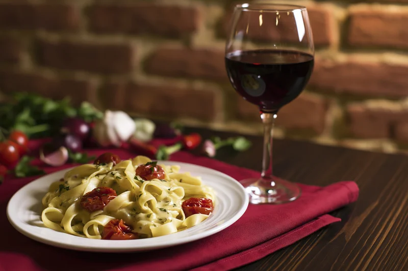 Plate of tagliatelle pasta with cherry tomatoes and herbs on a wooden table beside a glass of red wine