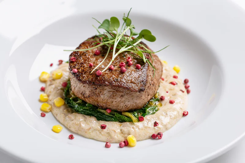 Seared beef steak on creamy puree with greens and garnish on white ceramic plate