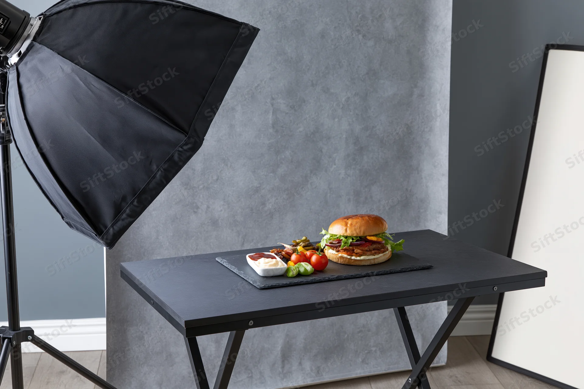 Burger with vegetables and sauce on slate plate on table in studio with softbox lighting and reflector
