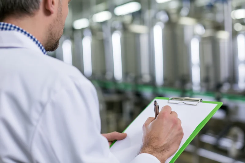 Technician in white lab coat writing on clipboard in factory setting