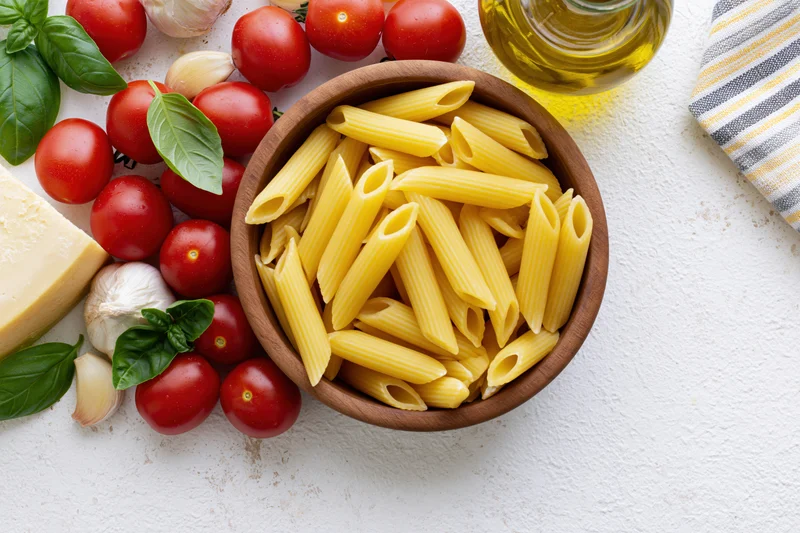 Raw penne pasta in a wooden bowl surrounded by cherry tomatoes, basil, garlic, cheese and olive oil on a white surface