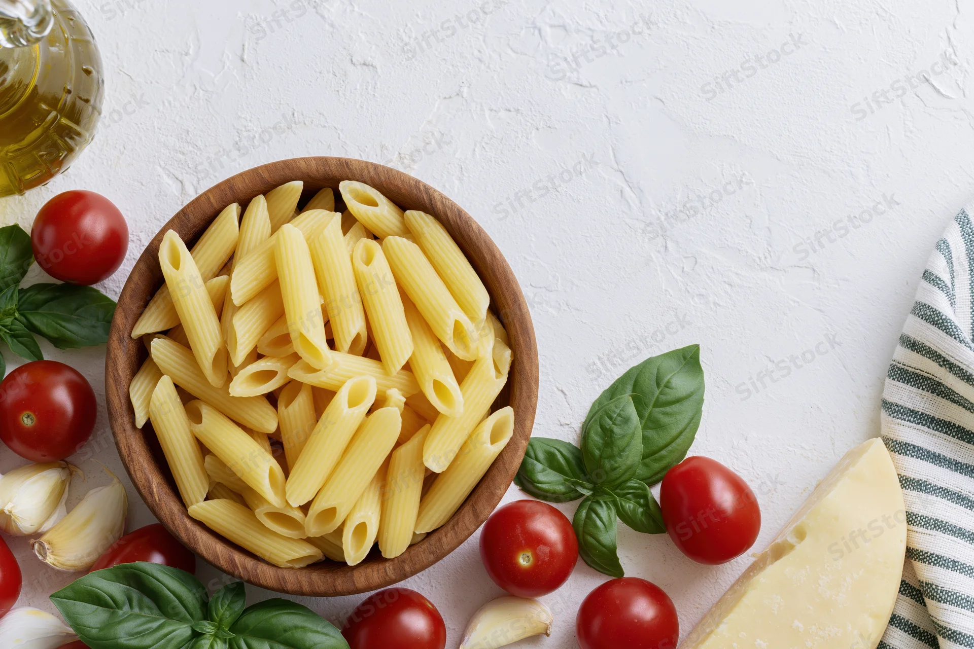 Raw penne pasta in a wooden bowl surrounded by cherry tomatoes, basil, garlic, cheese and olive oil on a white textured surface