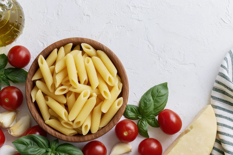 Raw penne pasta in a wooden bowl surrounded by cherry tomatoes, basil, garlic, cheese and olive oil on a white textured surface