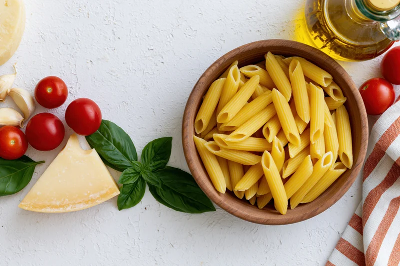 Raw penne pasta in a wooden bowl surrounded by cherry tomatoes, basil, parmesan, garlic, and olive oil on a white surface
