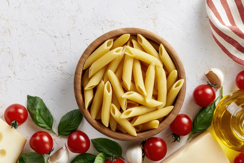 Raw penne pasta in a wooden bowl surrounded by cherry tomatoes, basil, garlic, cheese and olive oil on a white surface